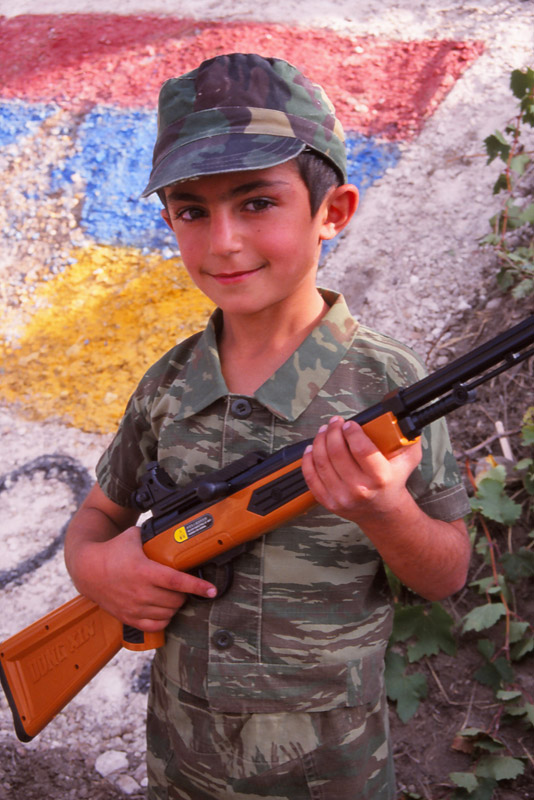 A boy armed with a plastic rifle stands guard over the Armenian flag in Areni