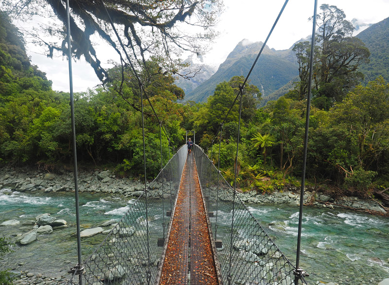 A suspension bridge crosses Hidden Falls Creek with the Darran Mountains in the distance